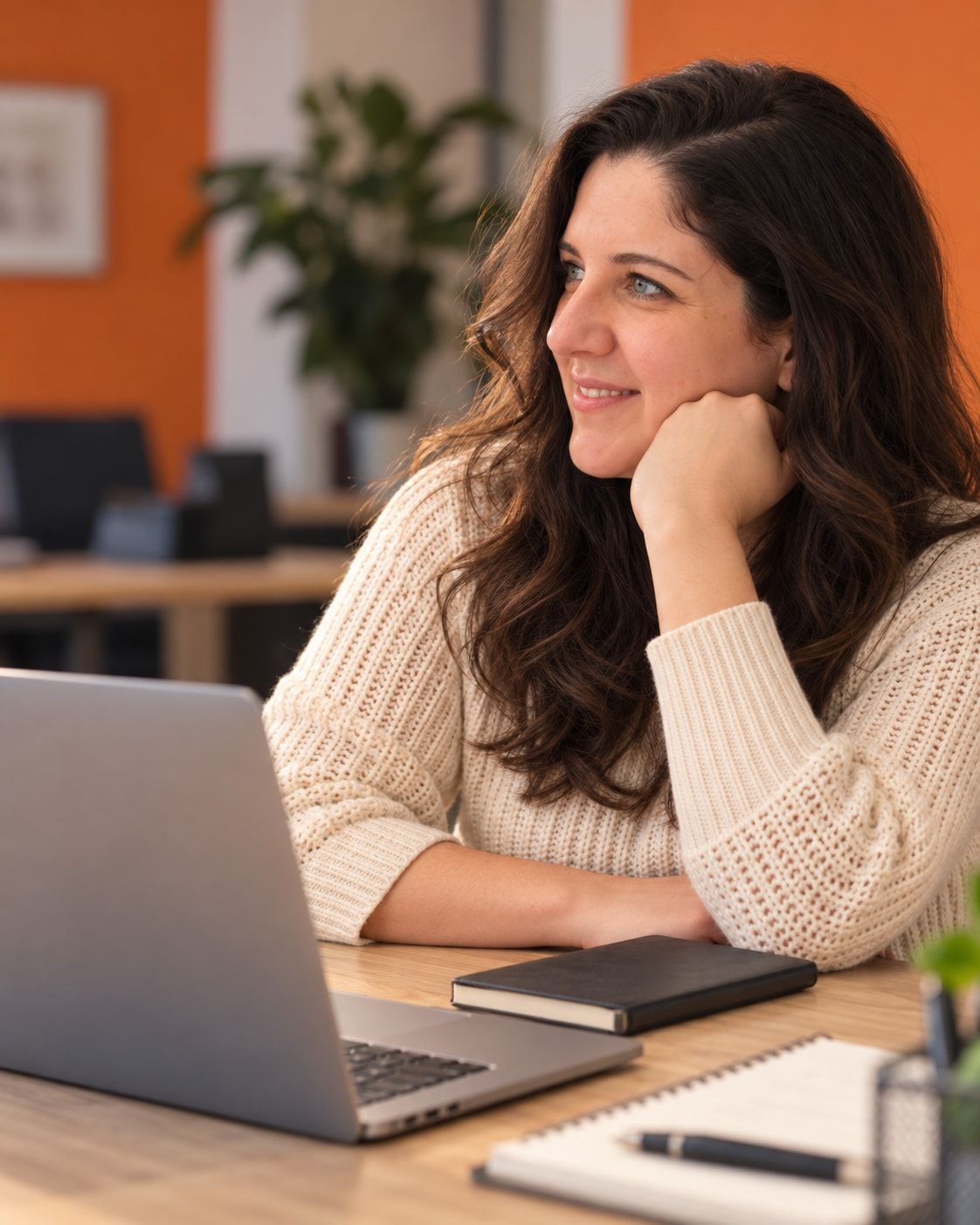 Anne Brussig sitzt im hellen Strickpullover am Schreibtisch vor einem Laptop, stützt das Kinn auf die Hand und schaut lächelnd zur Seite.