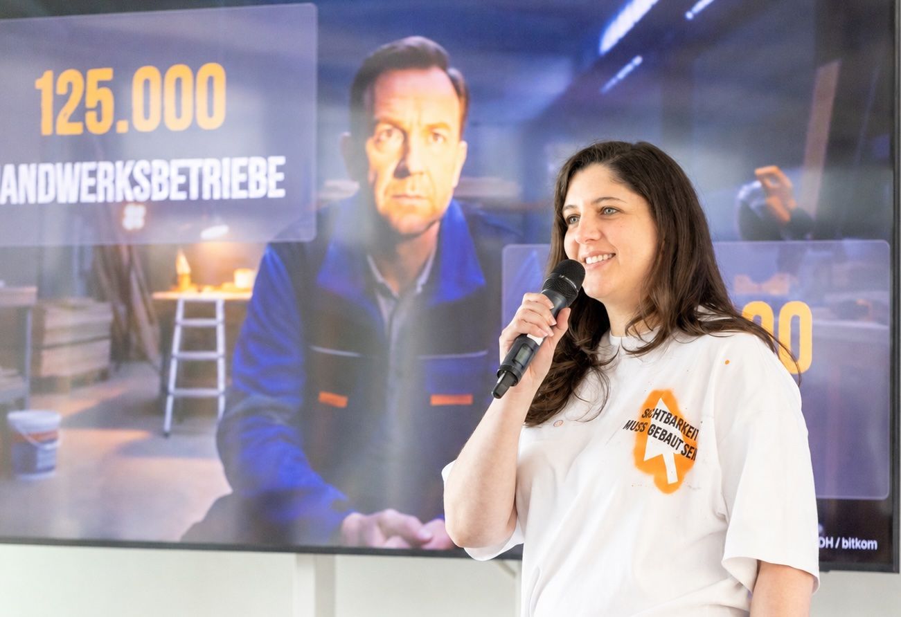 Anne Brussig spricht mit Mikrofon vor einer Leinwand mit der Folie „125.000 Handwerksbetriebe“, auf ihrem T-Shirt steht „Sichtbarkeit muss gebaut sein“.
