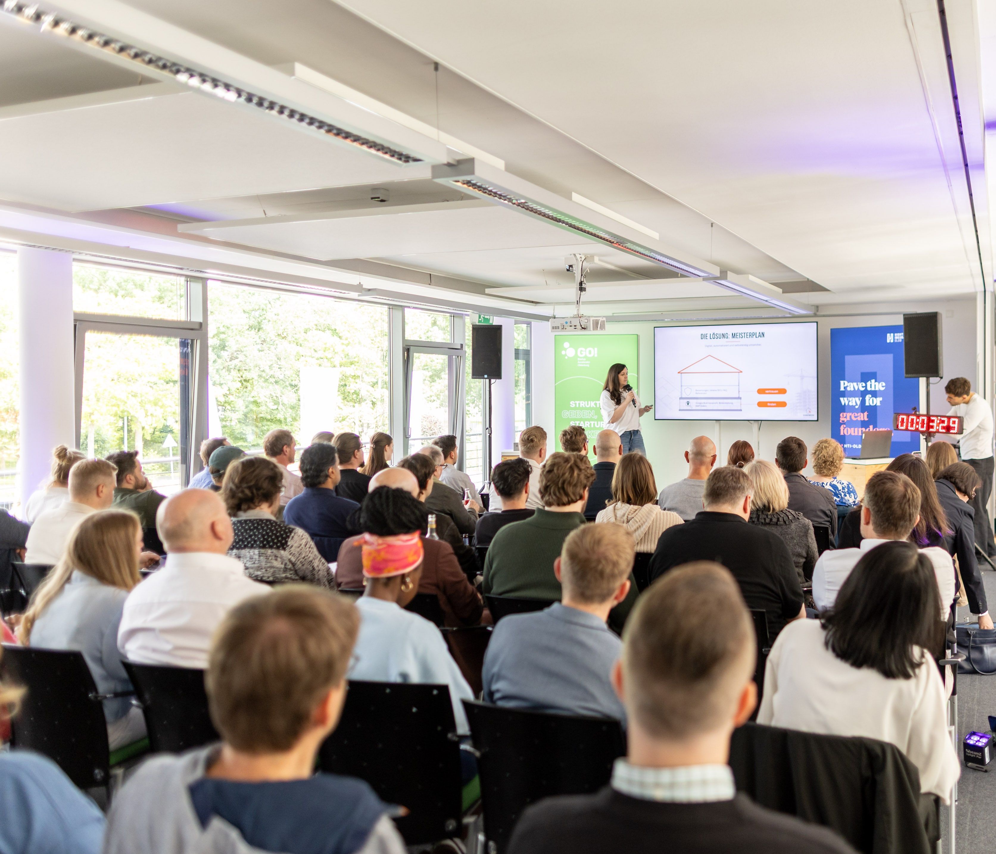 Blick über das voll besetzte Publikum beim Startup Spotlight Oldenburg auf Anne Brussig, die mit Mikrofon vor einem Bildschirm mit der Folie „Die Lösung: Meisterplan“ spricht.